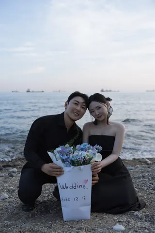 해변에서 파란 꽃다발과 결혼 날짜가 적힌 종이를 함께 든 신랑과 신부